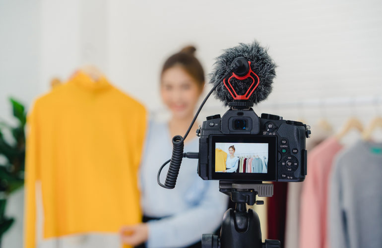 Asian fashion female blogger online influencer holding shopping bags and lots of clothes on clothes rack for recording new fashion video broadcast live video to social network by internet at home.